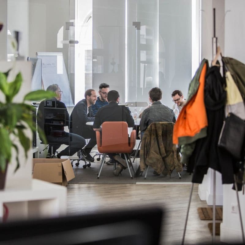 Kolleginnen und Kollegen der Logic Joe Group besprechen sich am Tisch hinter der Glaswand im hellen Altbau-Büro